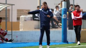 Soccer coach in a navy tracksuit on the sideline, gesturing with outstretched arms during a match.