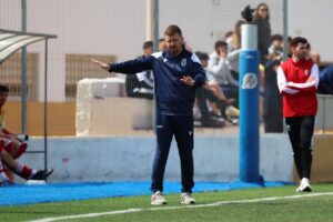 Soccer coach in navy tracksuit gesturing on the sideline during a game, with players on the bench behind a fence.