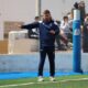 Soccer coach in navy tracksuit gesturing on the sideline during a game, with players on the bench behind a fence.