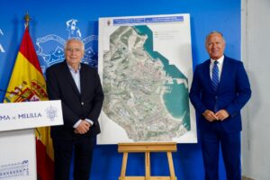 Two suited men stand beside a large urban planning map on an easel during an official presentation, with a Spanish flag nearby on a blue backdrop.