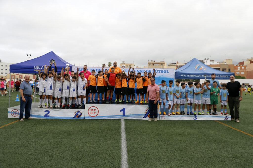 Youth soccer teams on a winners podium holding trophies, orange, white, and blue jerseys, with coaches nearby.