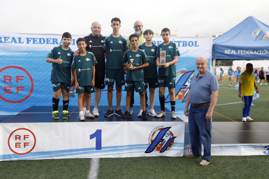 Youth soccer team on a first-place podium with coaches, holding trophies after a tournament sequence by the field side.