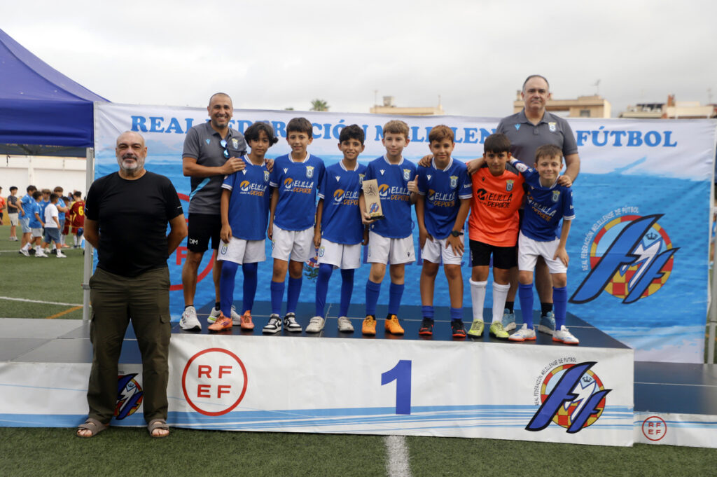 Youth soccer team on the first-place podium with two coaches, holding a trophy; blue uniforms and a football banner in the background.