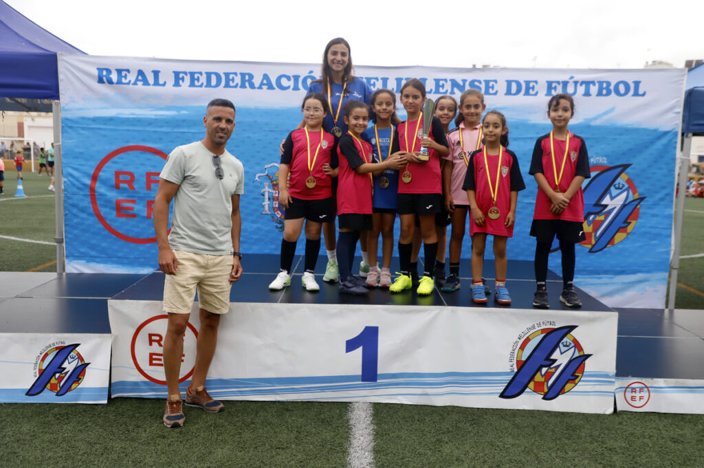 Young girls on a winners' podium with medals and a trophy, coached by a man, at a football event.