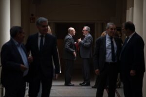 Two men in dark suits converse in a dim corridor, one gesturing with his hands and a red folder in view, while others linger nearby.