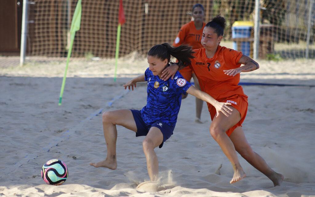 Two female beach soccer players compete for the colorful ball on a sandy field, one in blue and one in orange, with a goal net in the background.