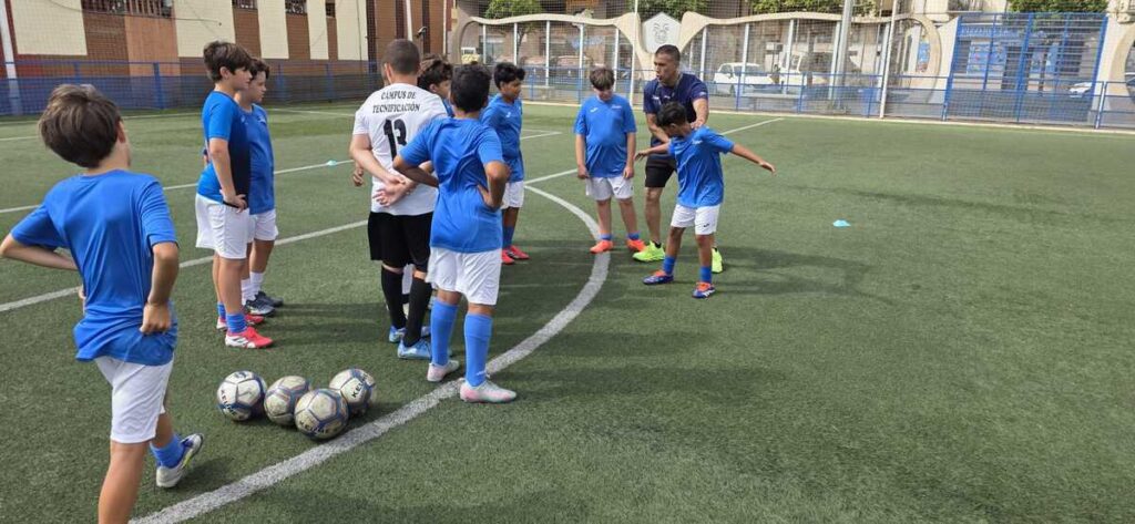 Soccer coach demonstrates drill to a line of young players in blue jerseys on an outdoor field with balls at their feet and cones nearby.