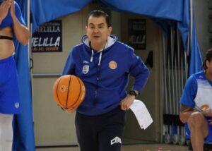 Basketball coach in a blue hoodie holding an orange basketball with a clipboard during practice.