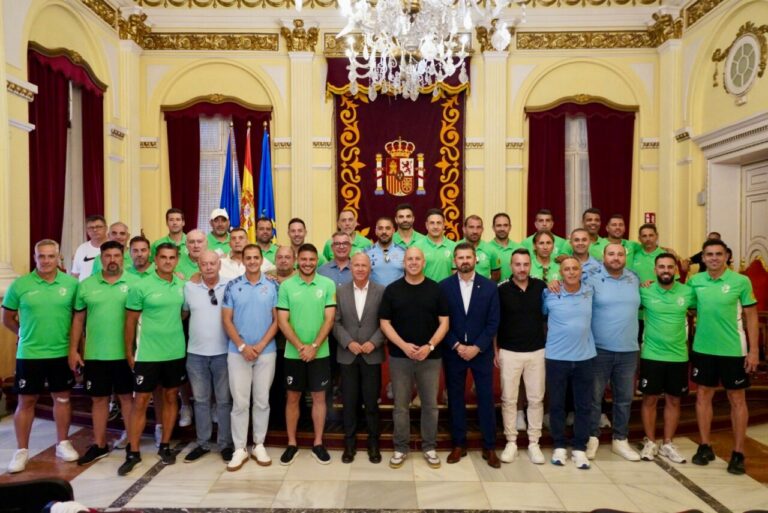 Group photo of a sports team and officials in a gilded ceremonial hall with flags and a royal crest banner behind them, all posing on steps.