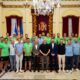Group photo of a sports team and officials in a gilded ceremonial hall with flags and a royal crest banner behind them, all posing on steps.
