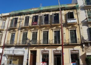 Three-story beige building with ornate window frames and iron balconies, above row of ground-floor shops on a sunny street