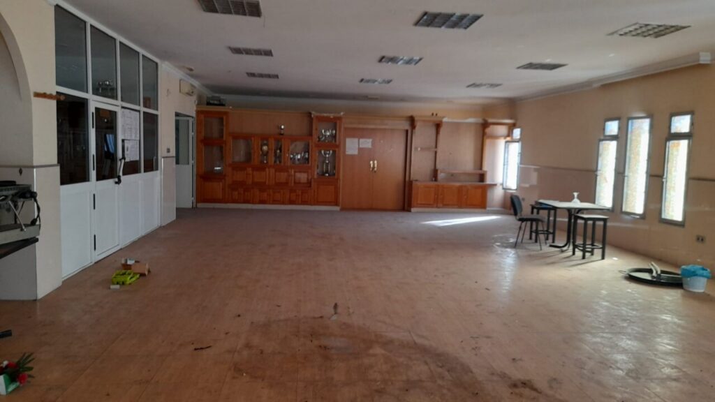 Spacious empty room with a dusty floor, wooden cabinets along the back wall, and a small table with stools near tall windows on the right.
