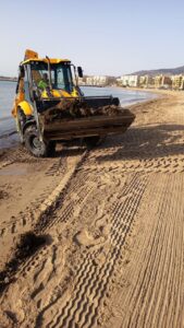 Yellow front-end loader on a sandy beach, scooping moist sand with city buildings in the background by the water's edge