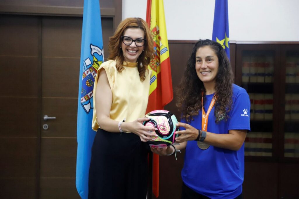 Two women standing indoors exchange a colorful soccer ball, with blue and red flags behind them and smiles on their faces.