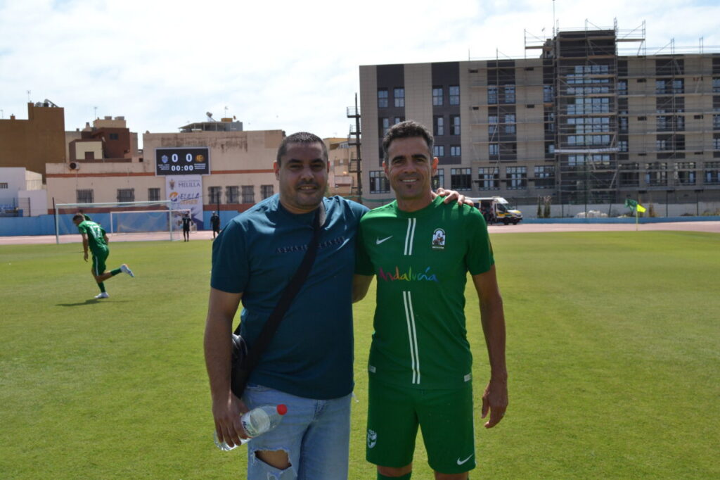 Two men pose together on a sunlit soccer field; one in a teal shirt with a shoulder bag, the other in a green soccer kit, both smiling.