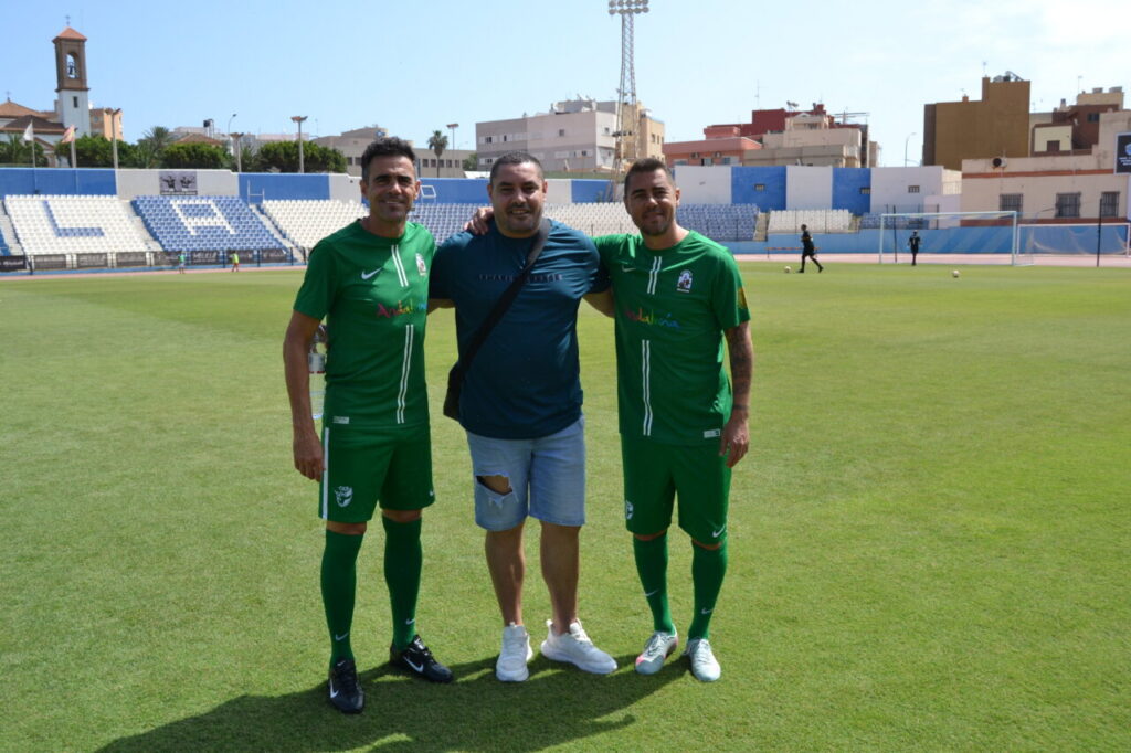 Three men pose together on a sunny football pitch; two wear green soccer kits, the third in a blue shirt and shorts with a crossbody bag around his shoulder.