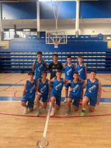 Ten young male basketball players in blue uniforms pose for a team photo on a gymnasium court, with a backboard and blue bleachers in the background.