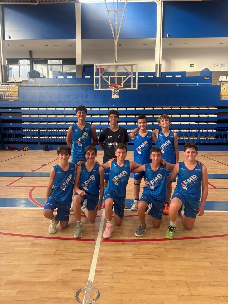 Ten young male basketball players in blue uniforms pose for a team photo on a gymnasium court, with a backboard and blue bleachers in the background.