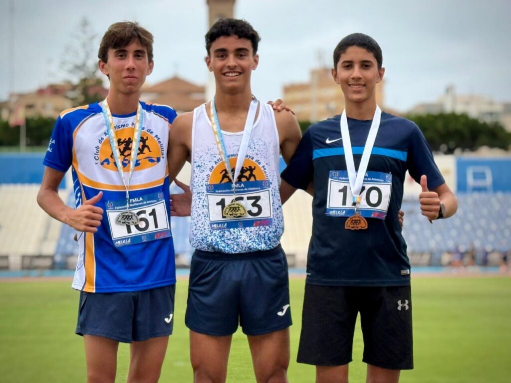 Three young male runners with medals pose on a track, smiling with arms around each other after a race, wearing blue and white outfits.