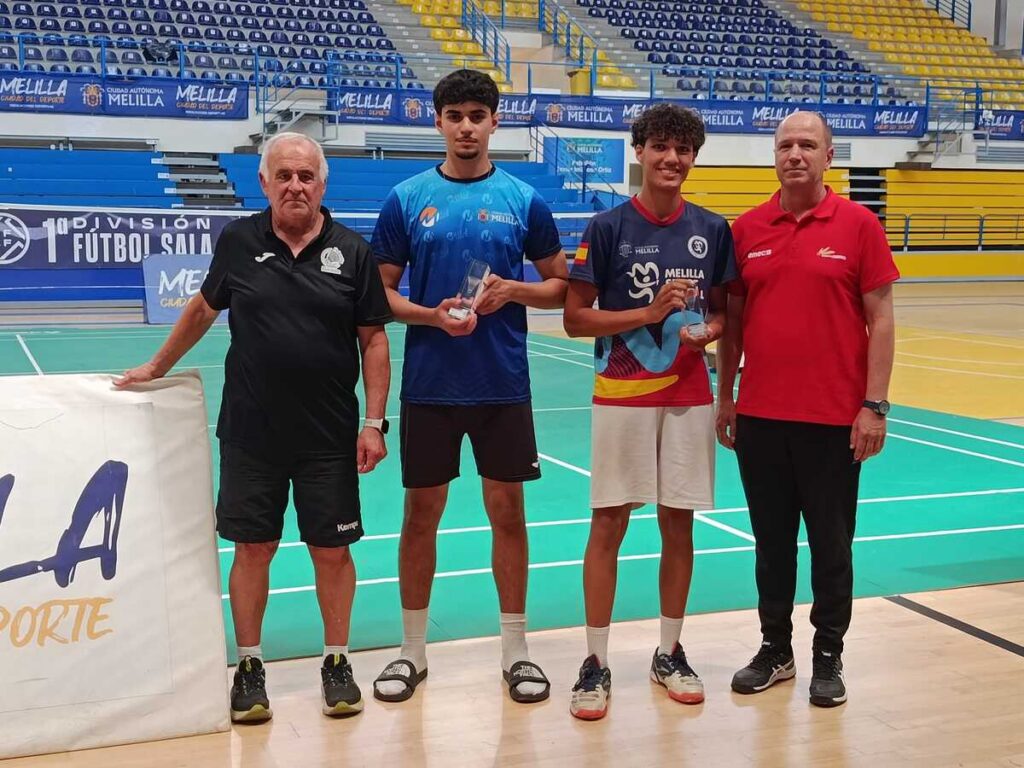 Four men pose on a futsal court; two young players in blue jerseys hold trophies, flanked by two coaches in black and red polos.