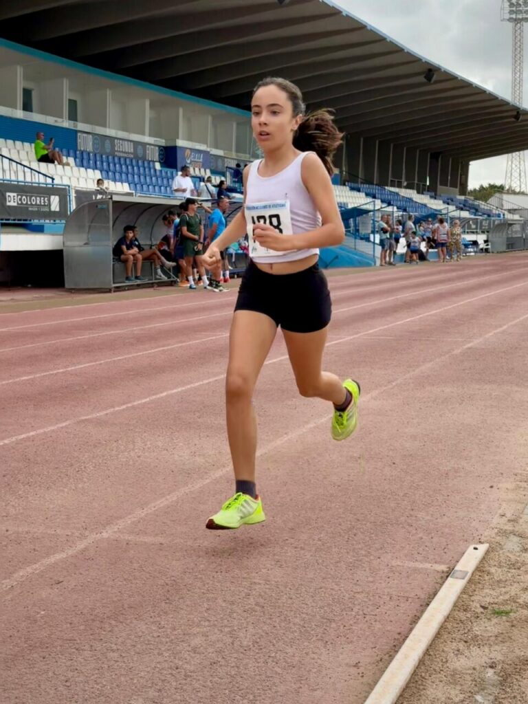 Young female track runner mid-stride on a red track in a stadium, wearing a white sleeveless top, black shorts, neon green running shoes, and a race bib on her chest.