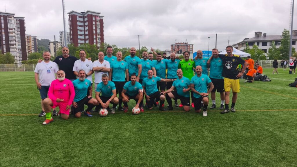 Group of soccer players in turquoise uniforms posing for a team photo on a green field, with urban buildings behind them.