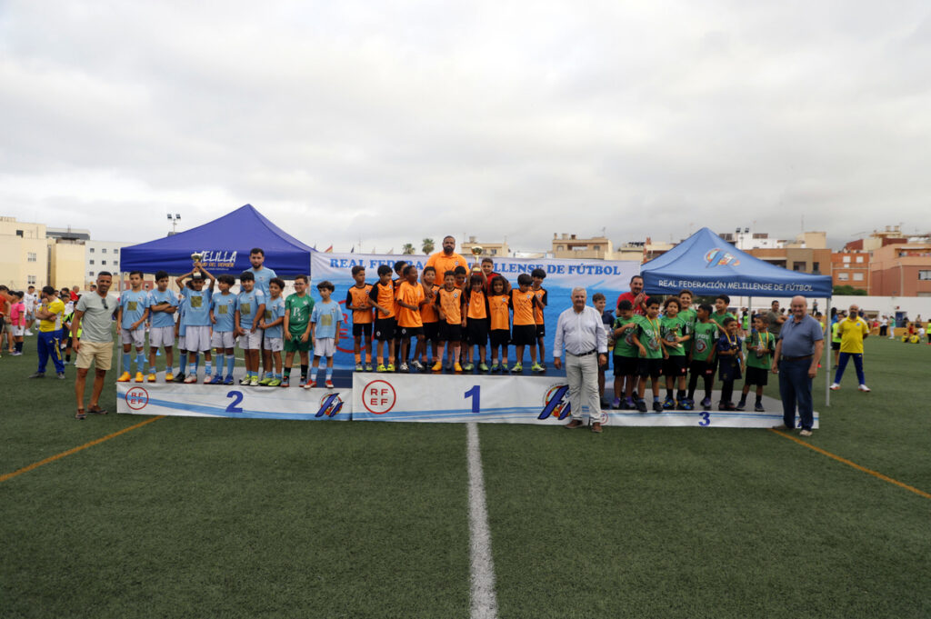 Youth soccer teams stand on a three-tier winners podium on a grassy field, wearing orange, blue, and green uniforms with tents in the background.