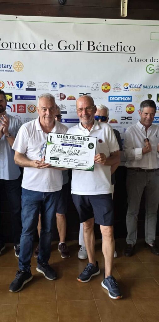 Two men in white polo shirts hold a large ceremonial check at a charity golf tournament, with a sponsor-filled backdrop behind them.