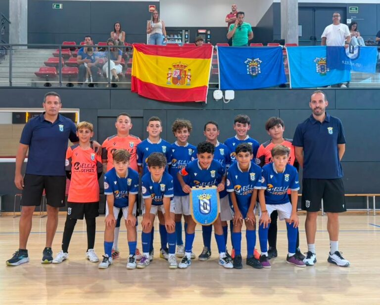 Youth soccer team posing indoors with two coaches, wearing blue uniforms, holding a shield, Spain flag and blue banners in the background.