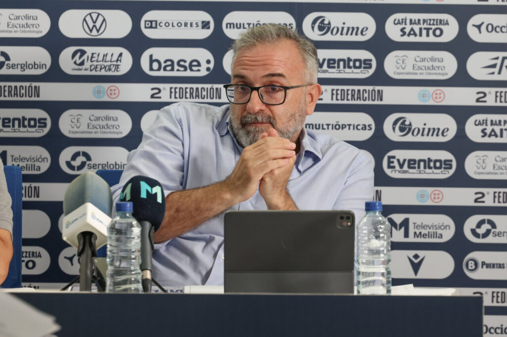 Man with glasses and gray beard speaks at a press conference, hands folded, microphones and a tablet in front, sponsor backdrop behind him.