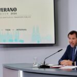 Man in a dark suit speaks at a conference desk with a microphone, a glass of water, and documents; a slide behind reads 'Verano Joven 2026' with 'Autobús — Tren — Interrail' and a Spanish flag nearby.