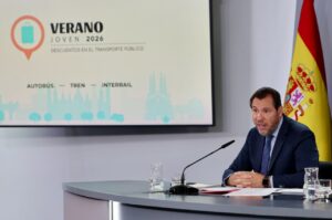 Man in a dark suit speaks at a conference desk with a microphone, a glass of water, and documents; a slide behind reads 'Verano Joven 2026' with 'Autobús — Tren — Interrail' and a Spanish flag nearby.