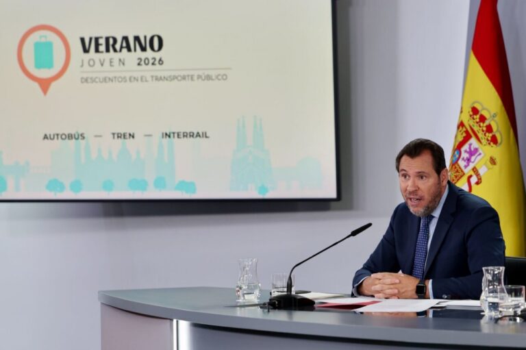 Man in a dark suit speaks at a conference desk with a microphone, a glass of water, and documents; a slide behind reads 'Verano Joven 2026' with 'Autobús — Tren — Interrail' and a Spanish flag nearby.
