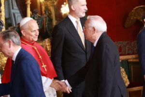 Two men in suits shake hands in a luxurious room; a cardinal in red robes stands nearby.