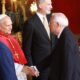 Two men in suits shake hands in a luxurious room; a cardinal in red robes stands nearby.