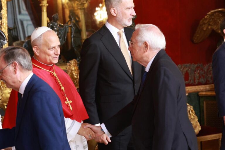 Two men in suits shake hands in a luxurious room; a cardinal in red robes stands nearby.