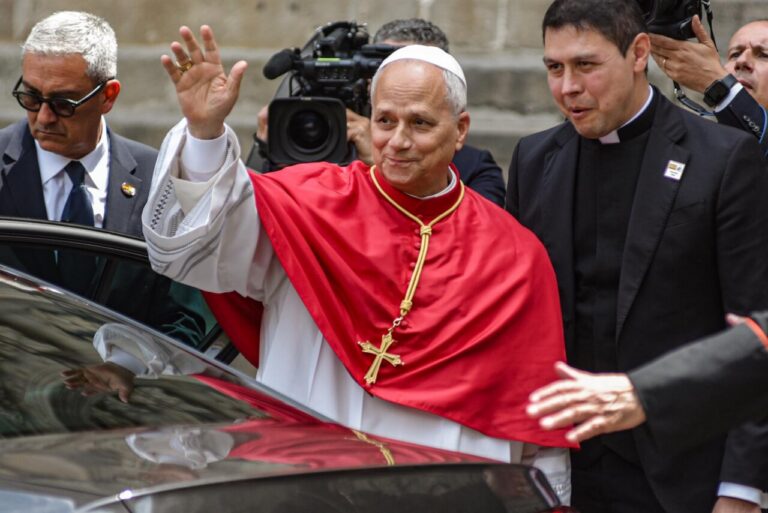 Clergyman in red ceremonial robes waves from a car doorway as security and reporters surround him.