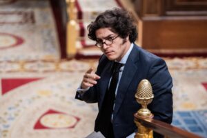 Man in a navy suit and glasses gesturing with his hand, standing beside a gilded banister in an ornate interior.