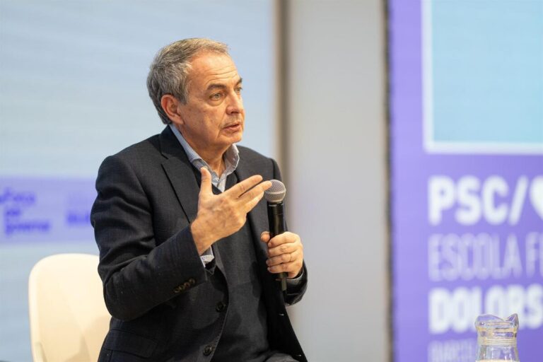 Middle-aged man in a dark blazer speaks into a handheld microphone, gesturing with his left hand during a panel or talk.