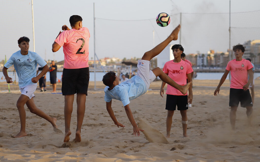 Group of boys playing beach soccer; a player in blue executes a bicycle kick as the ball flies overhead, others watch on a sandy field with nets in the background.