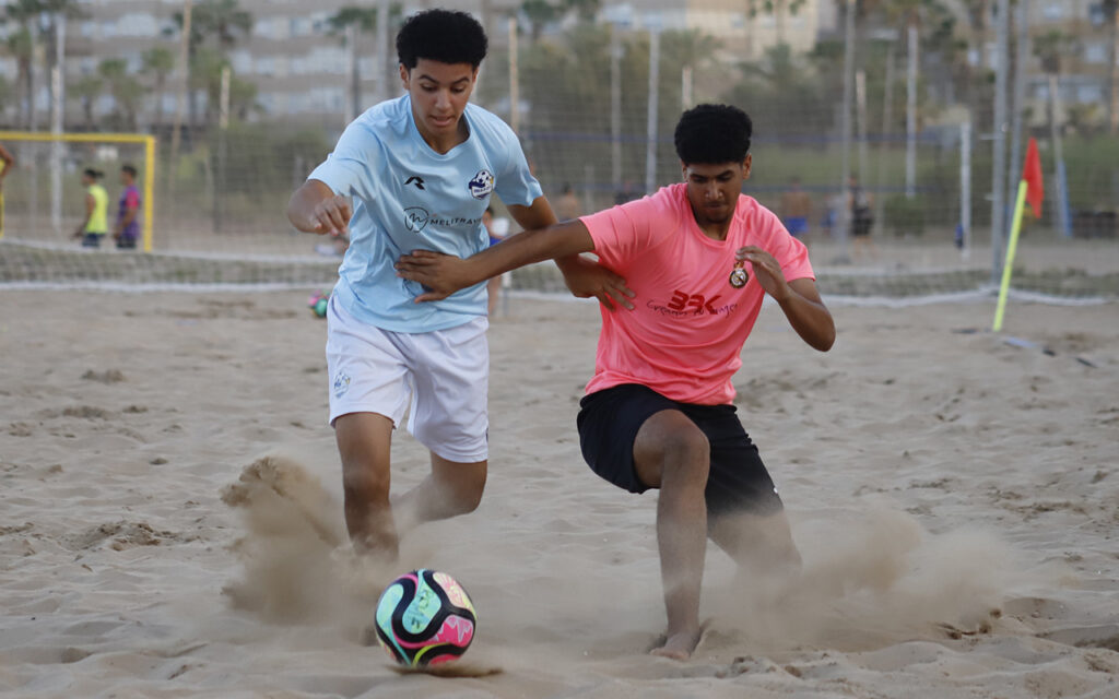 Two young men battle for a colorful soccer ball on a sandy beach soccer field, one in light blue, the other in pink.