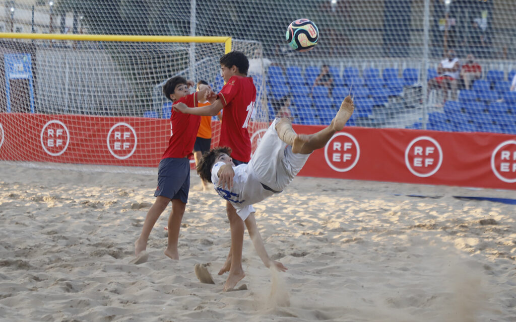 White-clad player performs a bicycle kick on a sandy beach soccer field as two red-shirted players challenge the ball near a goal, banners reading RF EF in the background.