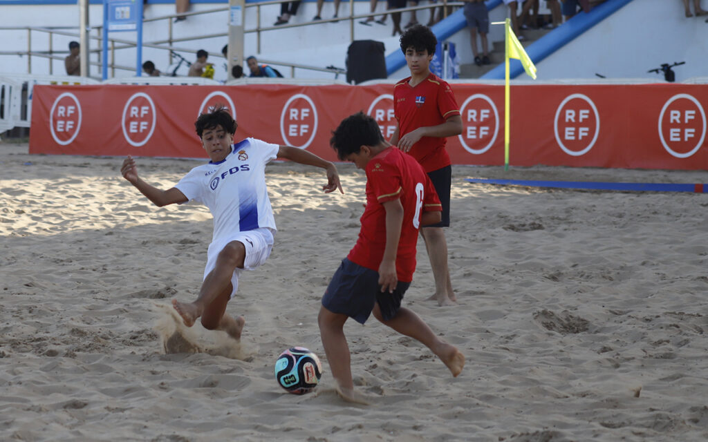 Three boys playing beach soccer on sandy surface, one in white kicking the ball while two in red chase nearby, with an orange barrier and spectator stands in the background.