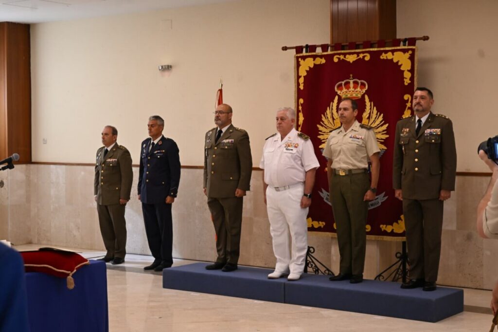 Six uniformed military officers stand in a line on a raised platform during a formal ceremony, with a decorative red banner behind them.