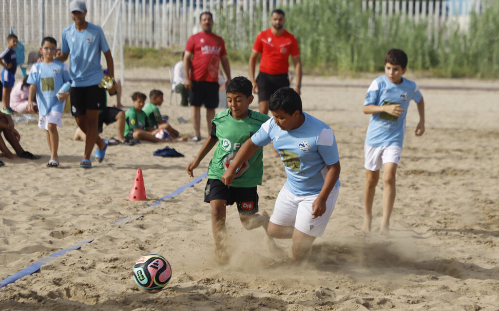 Youth soccer match on the beach; two boys in green and light blue shirts chase a colorful ball, kicking up sand.