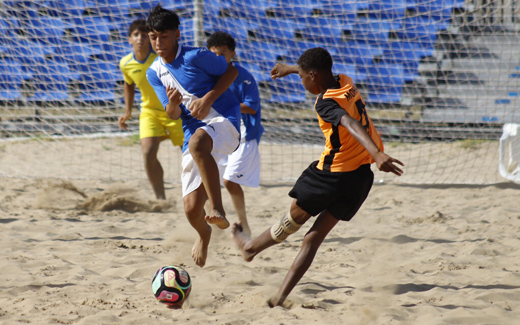 Beach soccer match with players contesting the ball on a sandy field near the goal net; blue and orange team uniforms visible in the scene.