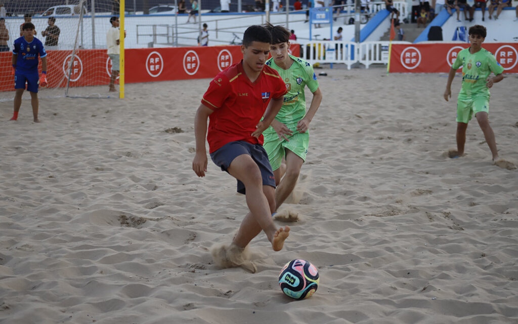 Young players compete in a beach soccer game; a boy in red kicks the colorful ball as a teammate in green closes in, sand flying.