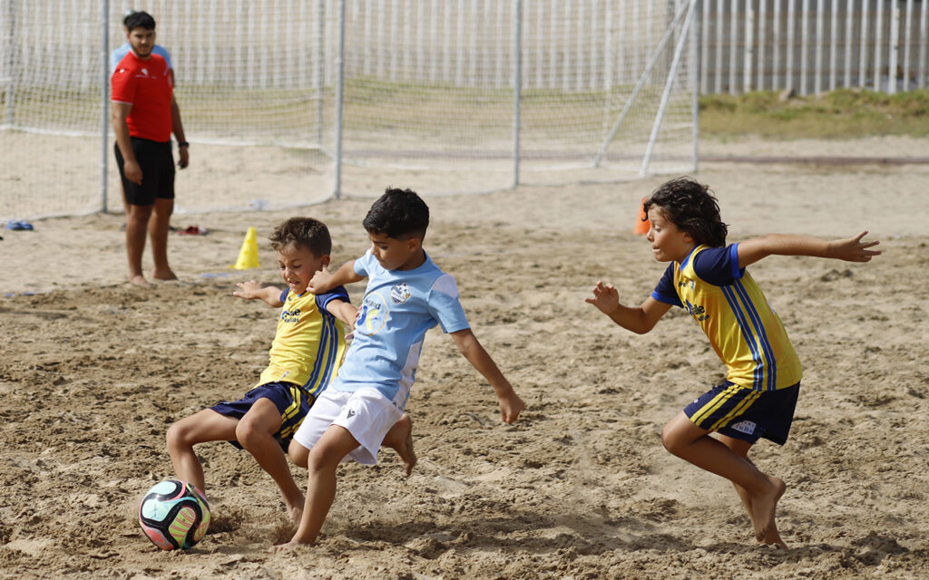 Three young boys compete for a soccer ball on a sandy field; two wear yellow jerseys, one in light blue shirt, all focused on the ball of the ground edge near a goal net behind them