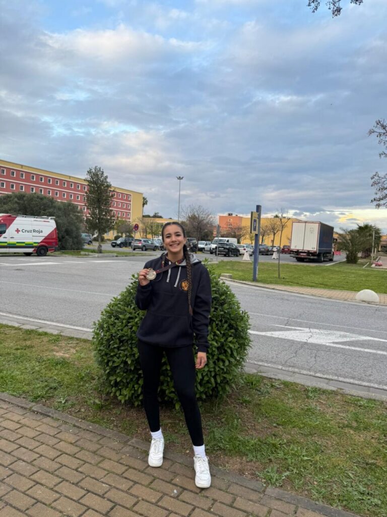 Young woman in a black hoodie with a crest, white sneakers, stands on a sidewalk in a parking lot, proudly holding a medal.