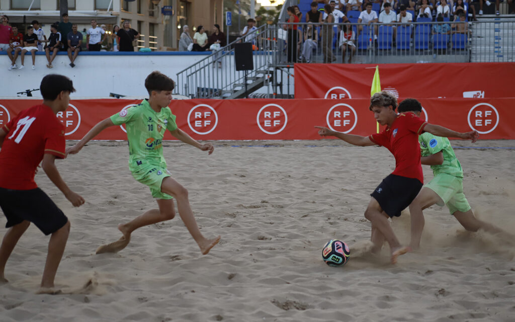 Young players in red and green jerseys compete for a beach soccer ball on a sandy field, with spectators and red banners in the background.